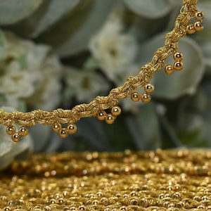 May include: Close-up of gold-colored trim with a braided design and small, dangling gold beads. The trim is likely used for embellishing clothing or crafts. The background is blurred, with a hint of white flowers.