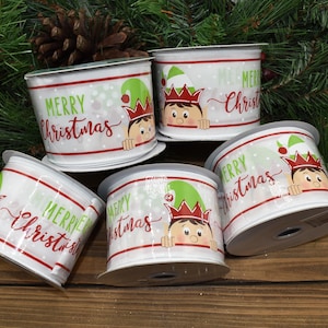 May include: White Christmas ribbon rolls with a red and green design. Each roll features the text "Merry Christmas" and a cartoon elf wearing a green hat and red crown. The ribbon has a dotted pattern.