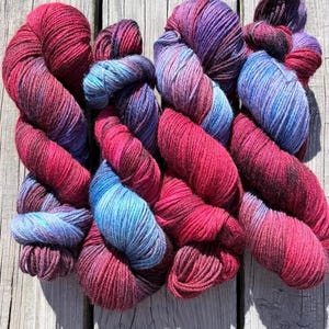 May include: Four skeins of hand-dyed yarn featuring a variegated color palette of deep magenta, vibrant fuchsia, bright sky blue, and dark violet. The yarn has a twisted, multi-ply texture with visible fiber strands throughout each individual skein.