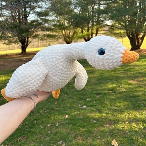 May include: A handmade white crochet goose plush toy with a long neck, blue eyes, and an orange beak. The goose has small orange feet and is held up against a green grassy background.