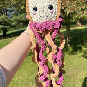 Puede incluir: Un peluche con forma de rebanada de pan con corteza color canela y centro crema, con ojos oscuros y una sonrisa bordada. Debajo de la rebanada de pan hay varios tentáculos largos y rizados en tonos alternos de hilo magenta y color canela.