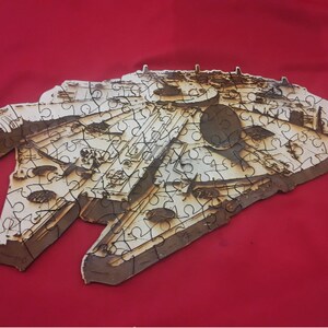 Puede incluir: Un rompecabezas de madera del Millennium Falcon, la nave espacial de Star Wars. El rompecabezas está ensamblado y muestra la nave icónica en detalle.