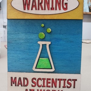 May include: A wooden sign with a yellow, blue, and white background. The sign has a red oval with the word "WARNING" in white letters. The sign also has a green beaker with bubbles and the text "MAD SCIENTIST AT WORK" in red letters.