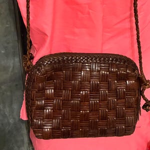May include: A brown woven leather crossbody bag with a long, braided strap. The bag has a rectangular shape with rounded edges. The background is a bright pink garment and a weathered, dark metal door.