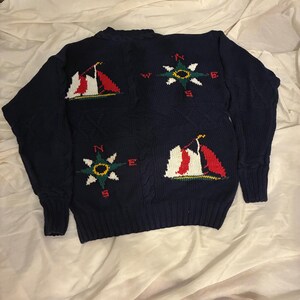 May include: Navy blue knit sweater featuring nautical designs. The sweater has a crew neck and long sleeves. It features embroidered sailboats and compass rose designs in red, white, green, and yellow.