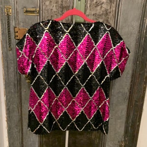 May include: A black and pink sequined top with a diamond pattern. The top has short sleeves and a scoop neck.
