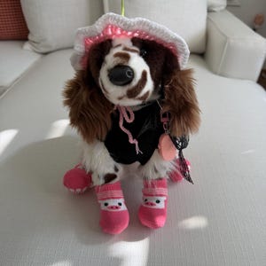 Peut inclure: Une peluche de chien épagneul portant un chapeau crocheté blanc et rose avec une petite fleur sur le dessus, un gilet noir avec un cordon rose et des chaussettes roses avec un motif de visage de cochon blanc. Le jouet a des poils marron et blancs.