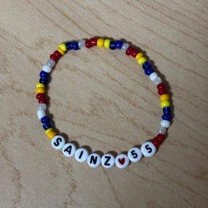 Könnte beinhalten: Ein Perlenarmband mit dem Namen "SAINZ 55" in weißen Buchstabenperlen. Das Armband hat ein Muster aus roten, gelben, blauen und weißen Perlen. Zwischen dem "Z" und der "55" befindet sich eine kleine rote Herzperle.
