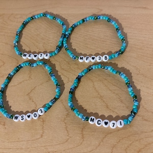 Könnte beinhalten: Vier Armbänder aus blauen und grünen Perlen. Jedes Armband hat weiße Buchstabenperlen, die Initialen und Zahlen buchstabieren, mit herzförmigen Perlen. Die Armbänder werden auf einer hellbraunen Holzoberfläche präsentiert.