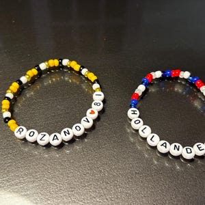 Peut inclure: Deux bracelets de perles. L'un est composé de perles jaunes, noires et blanches avec le nom "ROZANOV" et le chiffre "18". L'autre bracelet est composé de perles rouges, blanches et bleues avec le nom "HOLLANDER" et le chiffre "24".