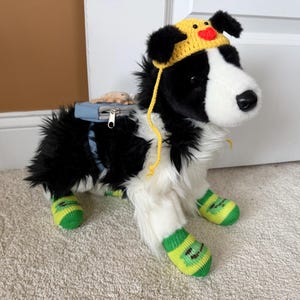 May include: A plush black and white dog toy wearing a yellow crocheted hat with a red heart detail, a light blue backpack with a silver zipper, and four green and yellow striped knit socks on its paws, standing in a stationary position.