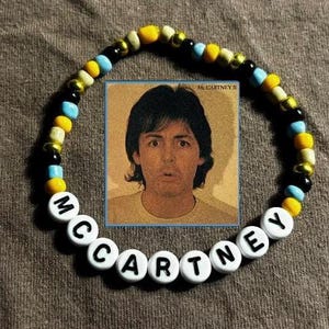 May include: A beaded bracelet with the name "MCCARTNEY" spelled out in white letter beads. The bracelet features a mix of yellow, black, and blue beads. A small image of a man is in the center of the bracelet.