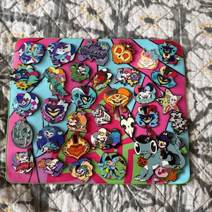 Könnte beinhalten: Eine Sammlung bunter Emaille-Anstecknadeln auf einem rosa-türkisfarbenen Hintergrund. Die Anstecknadeln zeigen verschiedene Zeichentrickfiguren und Designs, darunter einen Kürbis, einen Stern und ein Herz. Die Anstecknadeln sind auf einer rechteckigen Tafel angeordnet.