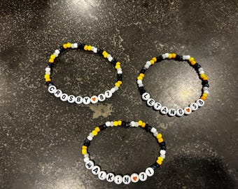 May include: Three beaded bracelets with black, white, and yellow beads. Each bracelet spells out a name and number with white letter beads. The bracelets say "CROSBY 87", "LETANG 58", and "MALKIN 71".