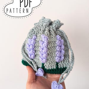 May include: A crocheted lavender-themed drawstring bag. The bag is gray with three lavender flower designs in purple. The base is green, and the drawstring is gray. The text "PDF PATTERN" is in a speech bubble.