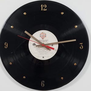 May include: A black vinyl record clock with gold-colored numerals and hands. The clock face features the label of "Grateful Dead The Best Of" album. The clock hands are gold with a red second hand. The numbers 3, 6, 9, and 12 are visible.