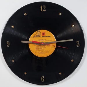 May include: A black vinyl record clock with gold-colored hour markers at 12, 3, 6, and 9 o'clock. The clock face features a yellow and orange label with the text "Fleetwood Mac" and "Reprise Records". The clock hands are silver and red.