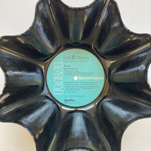 May include: A black, star-shaped bowl made from a recycled vinyl record. The center label is light blue with white text, including "Elvis" and "RCA." The bowl has a unique, wavy edge, and the record's grooves are visible.