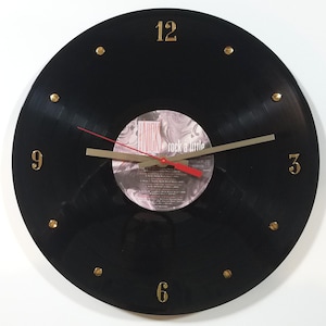 May include: A black vinyl record clock with gold-colored numerals at 12, 3, 6, and 9 o'clock, and gold-colored dots for the other hours. The clock hands are gold, with a red second hand. The center label reads "LINICK rock a little".