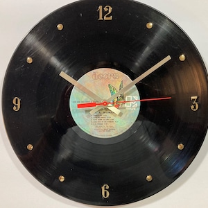 May include: A black vinyl record clock with gold-colored numerals and hands. The clock face features the album label for "L.A. Woman" by The Doors. The second hand is red. The clock is a unique decorative item.