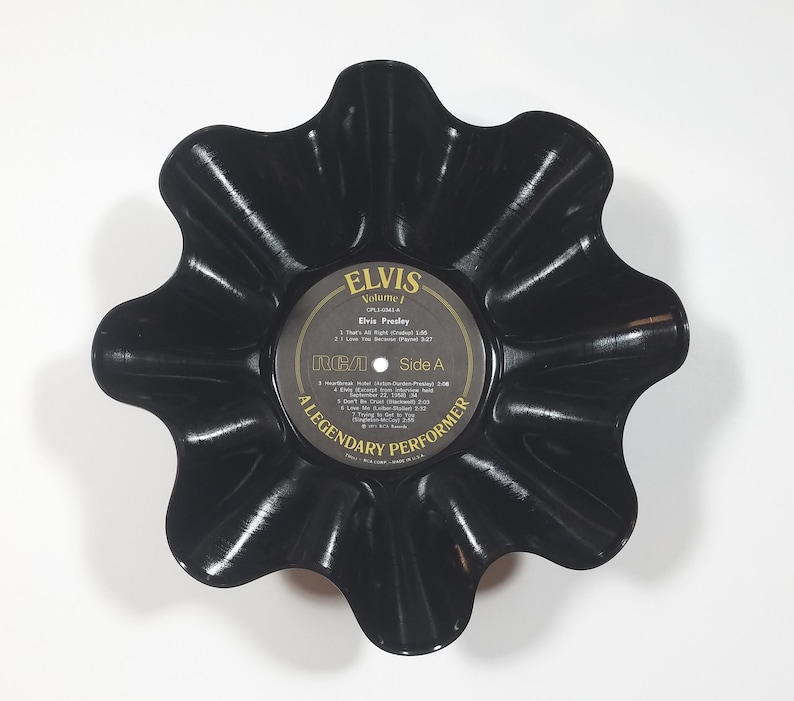 May include: A black, upcycled vinyl record bowl with a scalloped edge. The center label reads "ELVIS Volume 1" and features song titles. The bowl is a unique decorative item, perfect for holding small items or as a display piece.