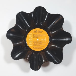 May include: A black, upcycled vinyl record bowl. The bowl is shaped like a flower with wavy edges. The original record label is visible, featuring text that reads "Elvis in Person at the International Hotel Las Vegas, Nevada."