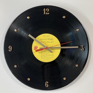 May include: A black vinyl record clock with gold-colored numerals at the 12, 3, 6, and 9 positions. The clock hands are silver and red. The record label is yellow with black text and the Rolling Stones logo.