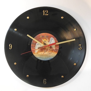 May include: A black vinyl record clock with gold numbers and a gold-colored minute hand. The center of the clock features a record label with the album art for Led Zeppelin's "Physical Graffiti" album. The album art features a winged figure with the text "Physical Graffiti" and "Led Zeppelin" printed on the label.