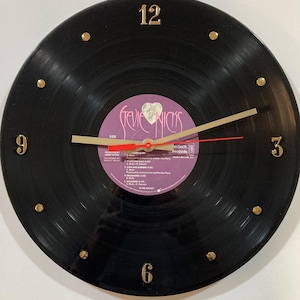 May include: A black vinyl record clock with gold-colored numerals and hands. The clock face features a purple label with the text "Stevie Nicks" and song titles. The hour markers are small gold studs.