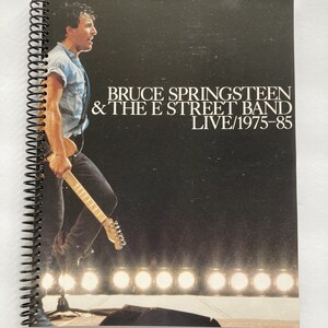May include: A black and white photo of Bruce Springsteen and the E Street Band on a concert stage. The photo is from a 1975-85 live album. Springsteen is holding a guitar and is facing the left side of the image. The stage is lit with spotlights.