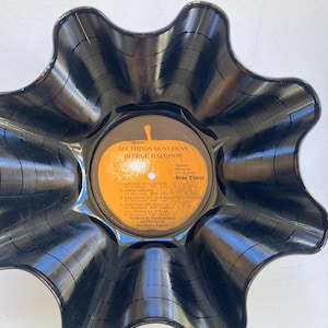 May include: A black, wavy-edged bowl made from a repurposed vinyl record. The center label is orange and features the text "ALL THINGS MUST PASS GEORGE HARRISON" and song titles. The bowl's unique shape and glossy finish create a striking decorative piece.