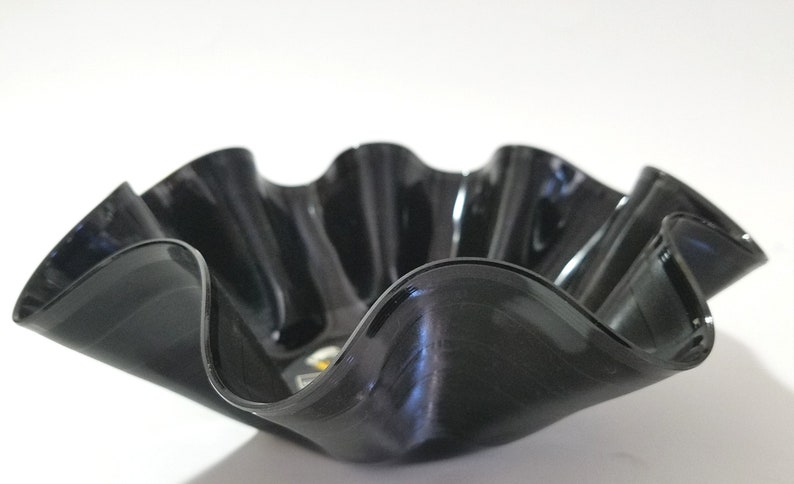 May include: A decorative bowl crafted from a recycled vinyl record. The bowl has a wavy, ruffled edge, and a glossy black finish. The bowl is a unique, eco-friendly home decor item.