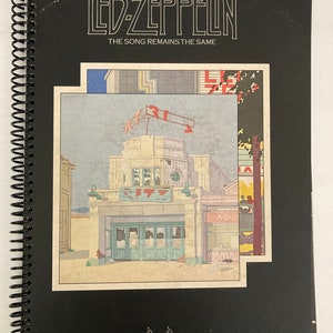 May include: A black spiral-bound book featuring the album art for Led Zeppelin's "The Song Remains the Same." The cover art depicts a building with a "RIBS" sign, with the band's name and album title at the top.
