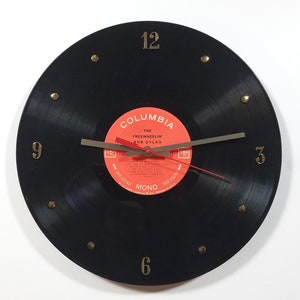 May include: A black vinyl record clock with gold-colored numerals and brass-colored studs. The clock face features a red and white label with the text "The Freewheelin' Bob Dylan". The clock hands are gray and red.