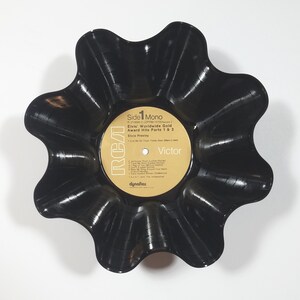 May include: A black, flower-shaped bowl made from a vintage vinyl record. The record label is visible, featuring text that reads "Elvis Worldwide Gold Award Hits Parts 1 & 2" and the RCA Victor logo. The bowl has a wavy, scalloped edge.