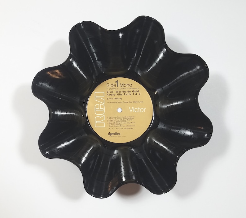 May include: A black, flower-shaped bowl made from a vintage vinyl record. The record label is visible, featuring text that reads "Elvis Worldwide Gold Award Hits Parts 1 & 2" and the RCA Victor logo. The bowl has a wavy, scalloped edge.