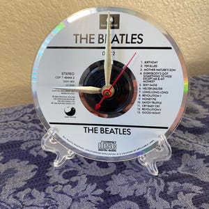 May include: A clock made from a compact disc featuring The Beatles album. The clock face displays the album title and tracklist, with silver hands and a red second hand. It is mounted on a clear plastic stand.