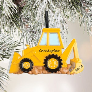 May include: A yellow construction vehicle Christmas ornament featuring a front blade and rear excavator arm. The side of the vehicle is printed with the name Christopher and the year 2023. It has black wheels and sits on a brown textured base.