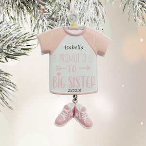 May include: Pink and white Christmas ornament shaped like a t-shirt with the text "Promoted to Big Sister" and the year "2023". The ornament also has a pink name tag with the name "Isabella" and pink shoes hanging from the bottom.