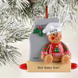 May include: Personalized Christmas ornament featuring a gingerbread person wearing a red and green sweater and a chef's hat. The ornament is decorated with a rolling pin and the text "Best Baker Ever!" and "2023". The gingerbread person's name is "Emily".