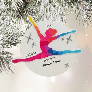 May include: A white, round Christmas ornament featuring a colorful dancer in a split. The dancer is in shades of red, pink, purple, blue, and yellow. The ornament has the text "2023", "Isabella", and "Atherton Dance Team".