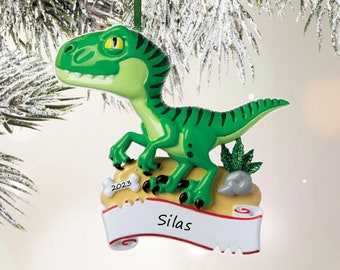 May include: A green dinosaur ornament featuring a yellow base with a white bone labeled 2023 and a white scroll banner with the name Silas printed in black. The dinosaur has dark green stripes, yellow eyes, and stands on a tan surface with a small green plant.
