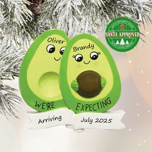 May include: A Christmas ornament shaped like two avocados, one with the name "Oliver" and the other with the name "Brandy". The avocados are holding a small brown circle in the middle. The ornament has a banner that says "We're Arriving" and another banner that says "Expecting July 2025".