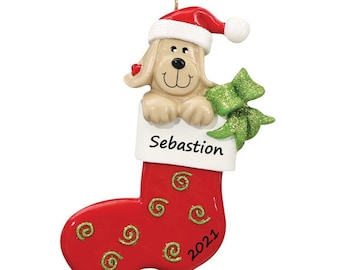 May include: A Christmas ornament featuring a tan dog wearing a Santa hat, peeking out of a red stocking. The stocking has green glitter swirls and the name "Sebastian" printed on it, with the year "2021" at the bottom. A green bow is on the side.