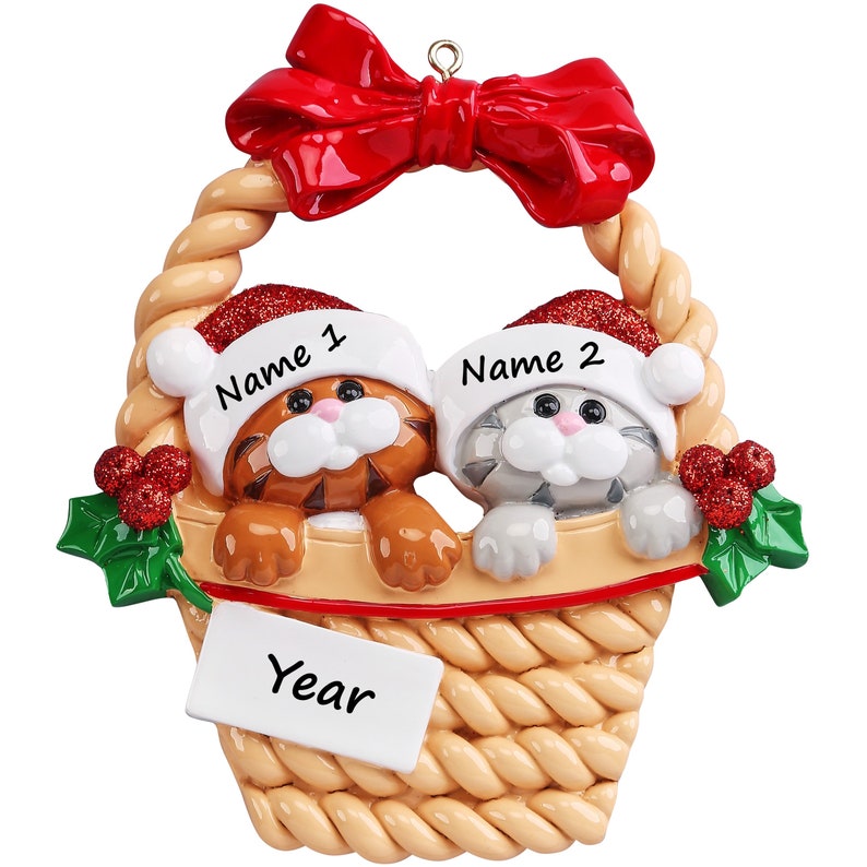 May include: A decorative hanging ornament featuring two kittens in a woven basket, topped with a red bow. The kittens wear glittery red Santa hats labeled Name 1 and Name 2. A white rectangular plaque at the bottom is labeled Year, with holly accents.