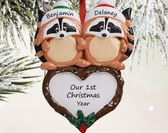 May include: A hanging Christmas ornament featuring two raccoons with black and white facial markings, wearing hats labeled Benjamin and Delaney. Below them is a heart-shaped frame with the text Our 1st Christmas Year and a small holly accent.
