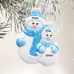 May include: A hanging Christmas ornament featuring two white snowmen figures wearing blue hats and scarves with snowflake patterns. The larger snowman is labeled Mom, and the smaller snowman is labeled Mila with the year 2026 printed below.