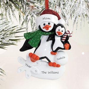May include: A Christmas tree ornament featuring two black and white penguins with orange beaks and feet. The larger penguin wears a red glittery hat and green polka-dot scarf. Text includes Mom, Isabelle, Year, and The Wilsons on the white base.