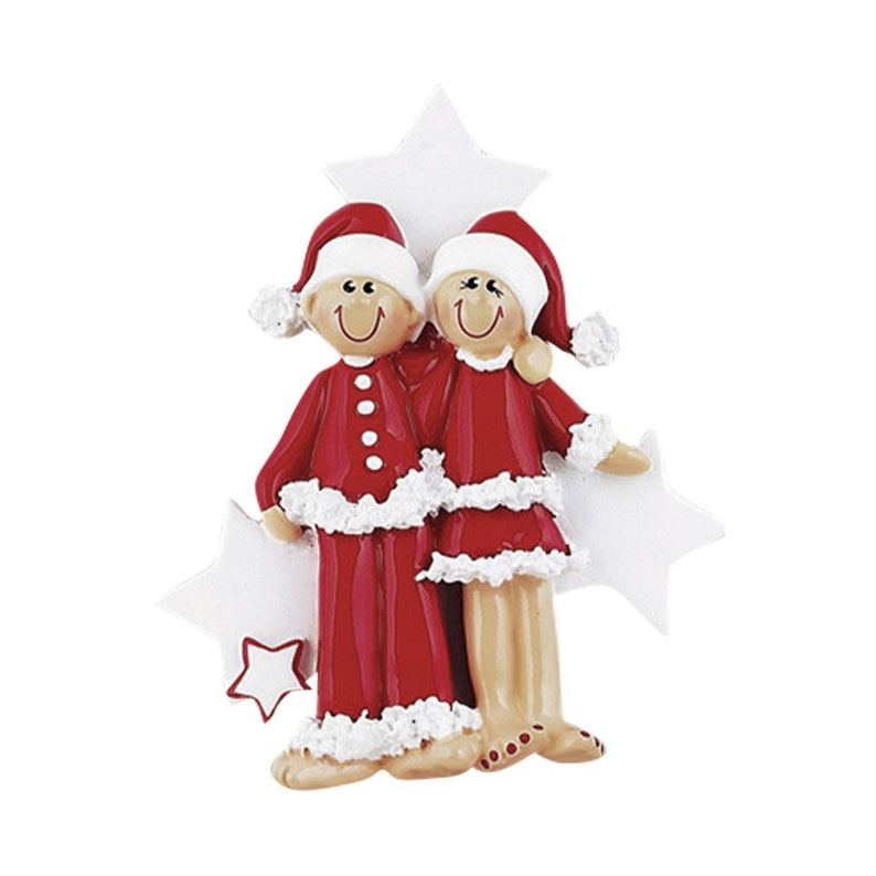 May include: A Christmas ornament featuring two figures standing side-by-side, both wearing red Santa hats and red outfits with white fuzzy trim. The figures have tan skin, smiling faces, and are positioned in front of three white star-shaped cutouts.