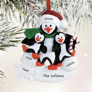 May include: A holiday ornament featuring three black and white penguins with orange beaks and feet. The largest penguin wears a red glittery Santa hat labeled Mom. The smaller penguins are labeled Noah and Isabelle. It includes a candy cane and a snowflake labeled Year and The Wilsons.